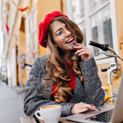 Cheerful female freelancer in glasses posing in outdoor cafe with computer and bicycle. Portrait of smiling curly woman in red beret drinking coffee on the street and working with laptop.