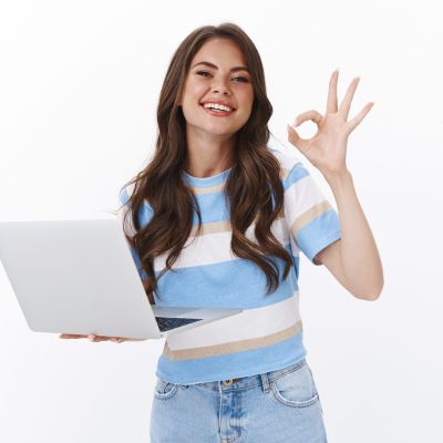 Confident carefree pretty young woman holding laptop, assuring she skilfull and professional smiling show okay ok sign, approve good article, accepting idea, stand white background satisfied.