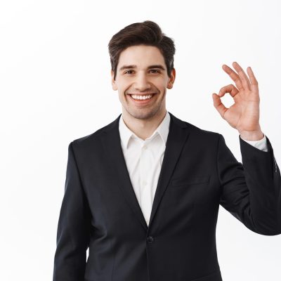 Handsome groom showing okay sign. Businessman in black suit make OK gesture and smiling satisfied, praise good job, well done, excellent gesture, white background.