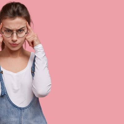 Image of displeased young lady keeps both index fingers on temples, suffers from migraine, wears round spectacles, being gloomy, isolated over pink studio wall with copy space on right side.
