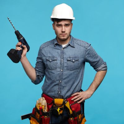 People, repair, equipment and renovation concept. Stylish young Caucasian repairman wearing hardhat, tool belt and overalls going to hang picture on wall, posing in studio with drill in his hand