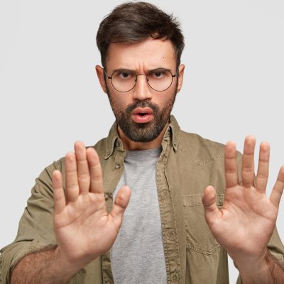 Photo of bearded young male shows stop gesture, has displeased facial expression, denies something, talks about forbidden things, wears fashionable shirt, isolated over white studio background