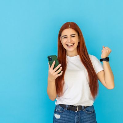 Portrait of beautiful woman looking at mobile phone and celebrating over blue background