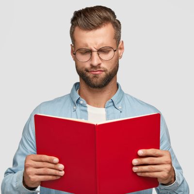 Serious clever young male teacher with trendy haircut, carries red book, tries to understand material, looks with indigant expression, wears round spectacles, stands against white background