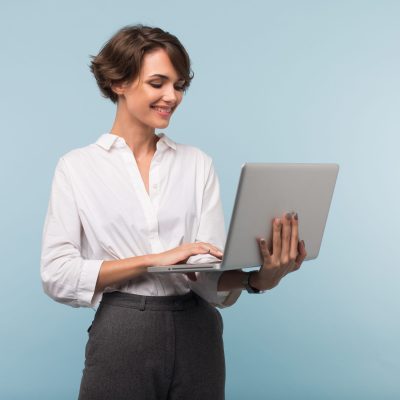 Young pretty smiling businesswoman with dark short hair in white shirt working on laptop over blue background