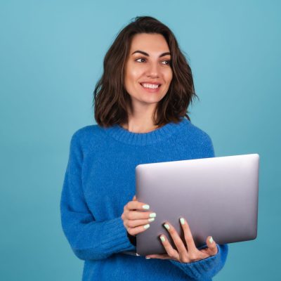 Young woman in a knitted sweater isolated holds a laptop, looks at the screen and laughs merrily